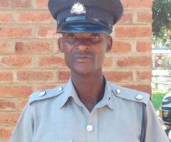 Male police officer in a light gray uniform and peaked cap standing against a brick wall.