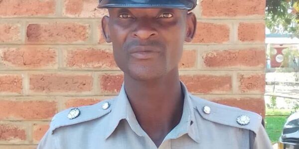Male police officer in a light gray uniform and peaked cap standing against a brick wall.