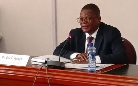 Man in a dark suit and glasses speaks into a microphone at a conference table with a water bottle and papers in front.