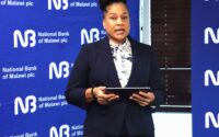 Woman in a navy blazer speaks at a National Bank of Malawi backdrop, holding a tablet in front of her.