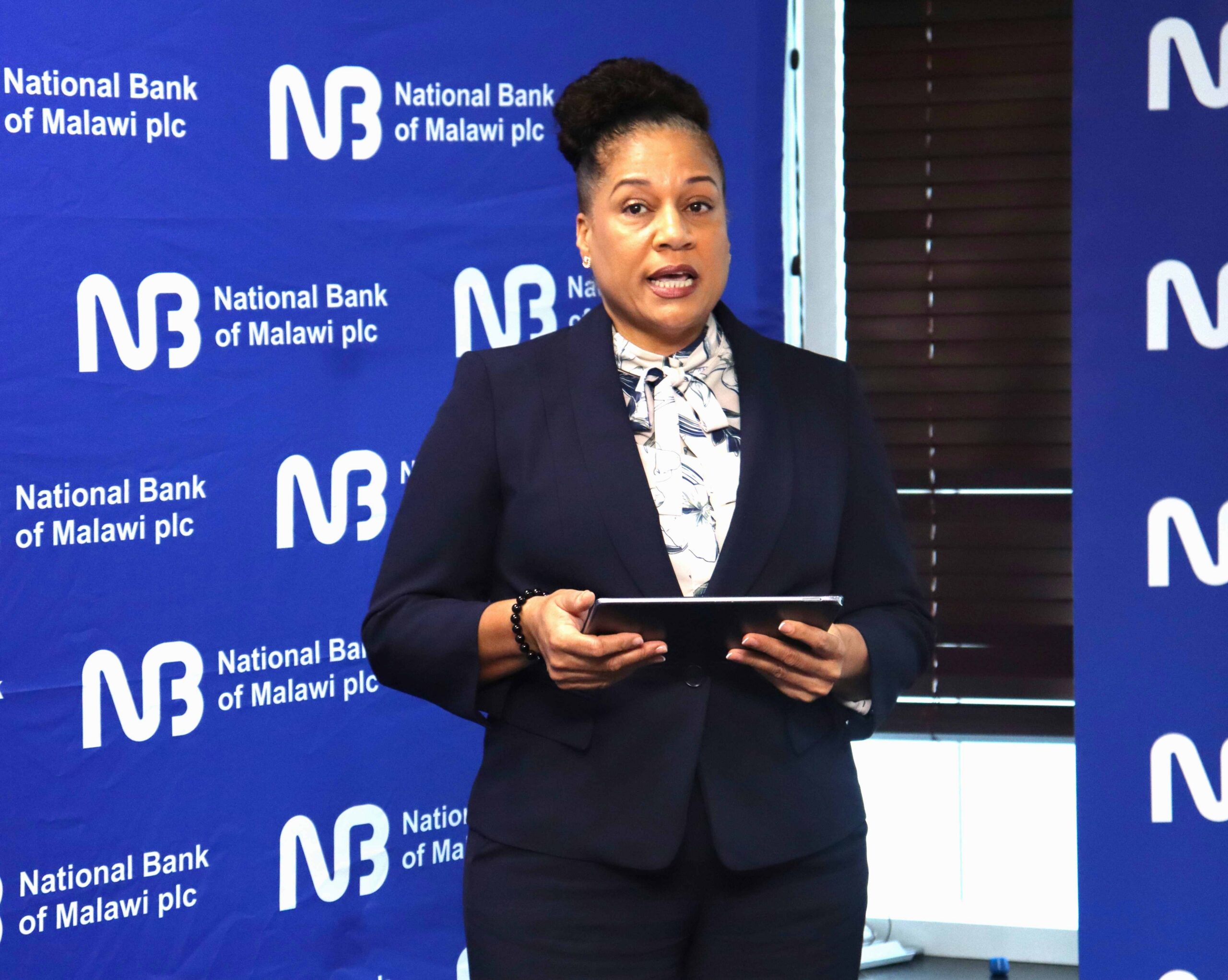 Woman in a navy blazer speaks at a National Bank of Malawi backdrop, holding a tablet in front of her.
