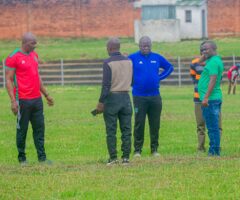 Group of men in athletic clothes standing on a grassy field, talking in a circle with a brick wall and small building in the background.