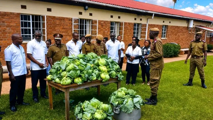 Group of uniformed officers and civilians gathered around a table piled with cabbages outdoors in front of a brick building.
