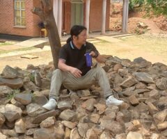Man wearing a black polo shirt and light pants sits on a pile of rocks outside a brick building, holding a blue water bottle and a cup.