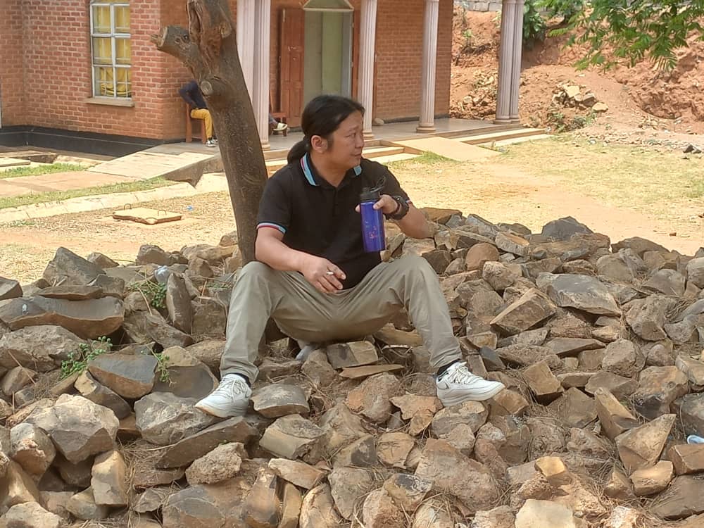 Man wearing a black polo shirt and light pants sits on a pile of rocks outside a brick building, holding a blue water bottle and a cup.